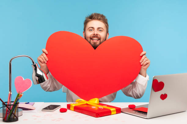 hombre barbudo feliz mirando detrás de gran corazón de papel rojo y sonriendo, sentado en el lugar de trabajo con portátil en corazones pegajosos, mostrando su amor - businessman 30s low key surprise fotografías e imágenes de stock