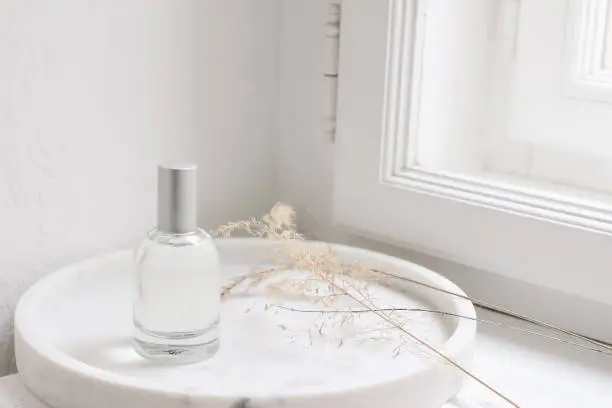 Feminine cosmetic still life scene. Glass bottle, flacon with perfume or eau de toilette and dry grass on white marble tray near window, selective focus, blurred window sill background.