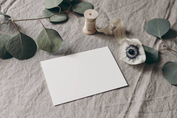 istock Wedding stationery mock-up scene. Blank greeting card on linen tablecloth background with white anemone flower, eucalyptus tree branches and silk ribbon. Feminine still life composition. High angle. 1295688319