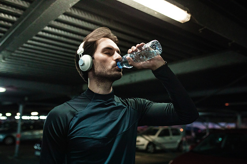 The young man is in an urban environment at night, he is wearing sports clothing and listening to music while drinking water