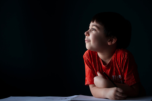 Cute little boy at his desk in front of the plain wall at home, looking outside and imagining what to draw in his drawing book