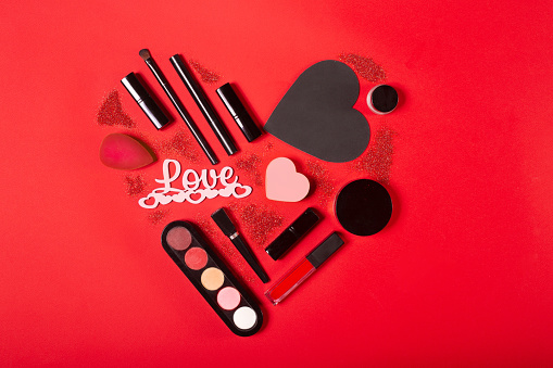 Cosmetic hear shape. Gift for girl on Valentines day. Love present for visagiste. Lipstick, eyeshadow, brush, eyeliner, sponge and hearts. on red background. Makeup flatlay