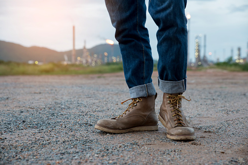 Closeup of the leg of engineers or safety officer, wearing blue jeans. safety brown shoes, standing in front of a refinery building.