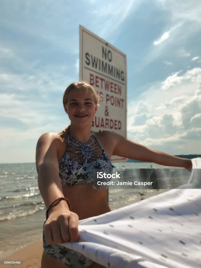 Happy teen girl with beach blanket on Lake Michigan. Pretty, smiling teenage girl with floral swimsuit on a beach near Lake Michigan, holding out blowing beach blanket and standing near a swim sign on the beautiful sunny beach enjoying the summer day. Taken in Gladstone Michigan, USA. Photography Stock Photo