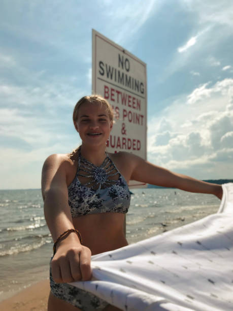 Happy teen girl with beach blanket on Lake Michigan. Pretty, smiling teenage girl with floral swimsuit on a beach near Lake Michigan, holding out blowing beach blanket and standing near a swim sign on the beautiful sunny beach enjoying the summer day. Taken in Gladstone Michigan, USA. gladstone michigan photos stock pictures, royalty-free photos & images