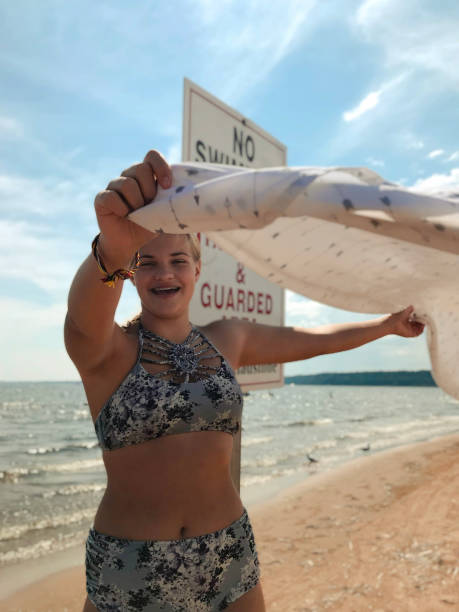 Happy teen girl with beach blanket on Lake Michigan. Pretty, smiling teenage girl with floral swimsuit on a beach near Lake Michigan, holding out blowing beach blanket and standing near a swim sign on the beautiful sunny beach enjoying the summer day. Taken in Gladstone Michigan, USA. gladstone michigan stock pictures, royalty-free photos & images
