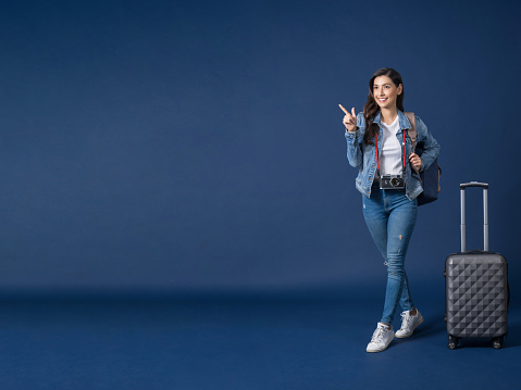 holiday and travel concept, Happy young Asian woman pointing and presenting on copy space  on blue color background, carry backpacker and baggage,