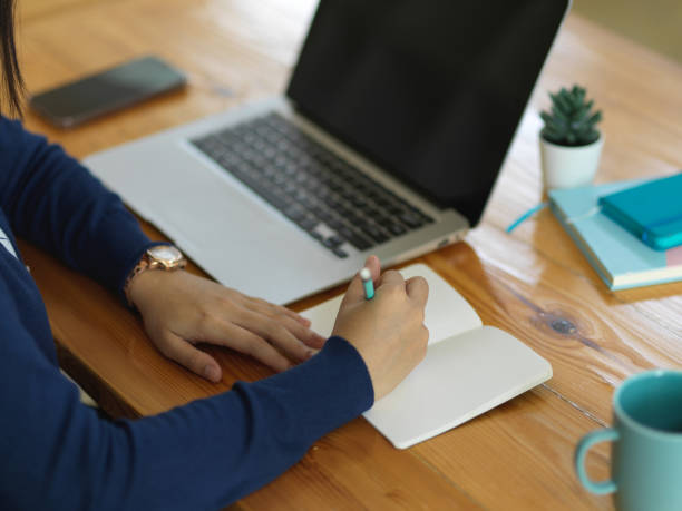 istock Female hand writing on blank notebook while working with laptop on wooden table 1295727965