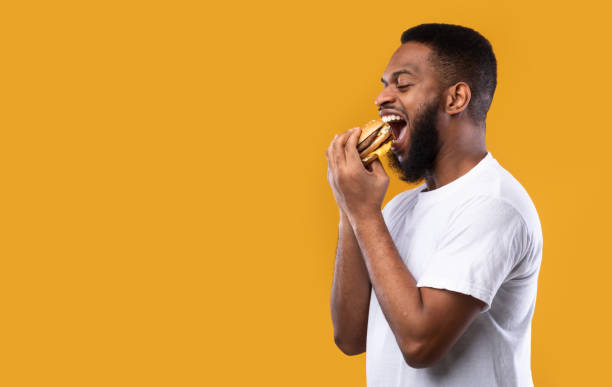 black millennial guy mordendo hambúrguer comendo sobre fundo amarelo, vista lateral - burger gourmet hamburger steak - fotografias e filmes do acervo