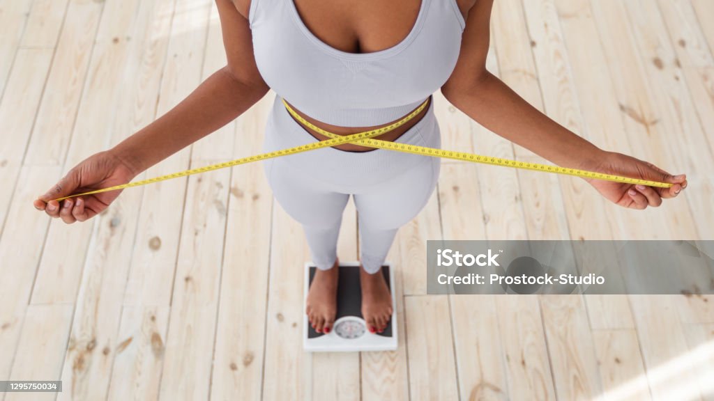Above view of African American lady measuring her waist with tape, standing on scales indoors, closeup Above view of African American lady measuring her waist with tape, standing on scales indoors, closeup. Young black woman showing results of slimming diet or liposuction, promoting healthy living Dieting Stock Photo