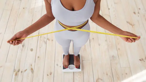 Photo of Above view of African American lady measuring her waist with tape, standing on scales indoors, closeup