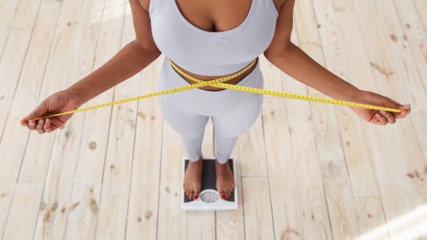 above view of african american lady measuring her waist with tape, standing on scales indoors, closeup - weight scale dieting weight loss imagens e fotografias de stock