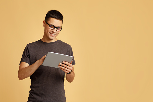 Work online, freelance, teacher and blogger. Smiling cute young male in brown t-shirt and glasses looks in digital tablet and surfing on internet, isolated on beige background, studio shot, free space