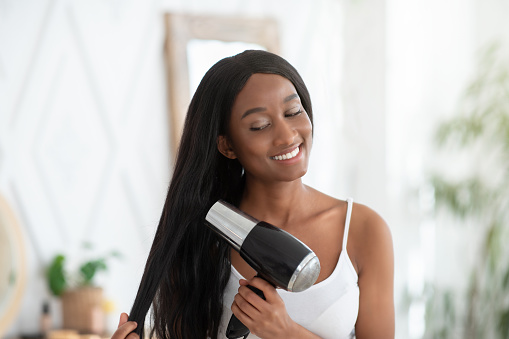 Wash hair, care and dry staying at home during covid-19 pandemic. Young happy beautiful african american lady doing hair styling with hair dryer at home in bedroom interior in morning, copy space