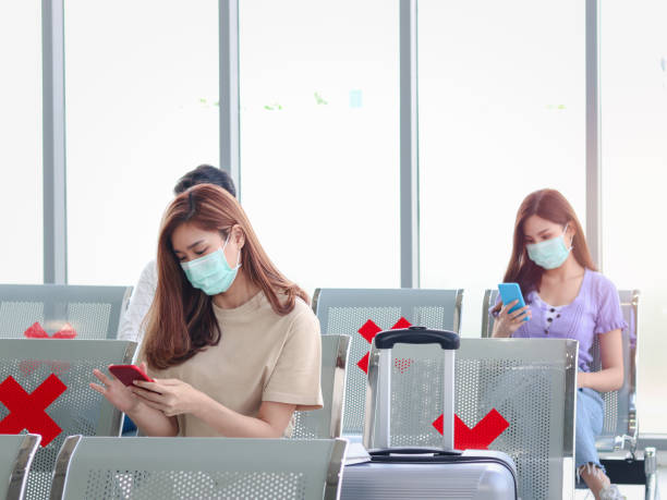 Asian young tourists wearing face mask keep sitting distance away from each other to prevent covid19 infection, using mobile phone, waiting airline flight at airport terminal, red cross shows on empty chair seat, social distancing and new normal travel concept. los turistas jóvenes asiáticos que usan máscara facial mantienen sentados lejos unos de otros para evitar la infección por covid19, utilizando el teléfono móvil, esperando el vuelo de la aerolínea en la terminal del aeropuerto, los espectáculos de