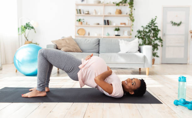 istock Black expectant lady doing abs exercises on mat at home, standing in half bridge pose, taking care of her future child 1295751168