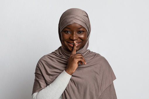 Mysterious Black Muslim Woman In Hijab Showing Shh Sign, Putting Finger On Lips, Smiling African Religious Lady In Headscarf Gesturing Hush And Keep Silence Over Light Studio Background, Free Space