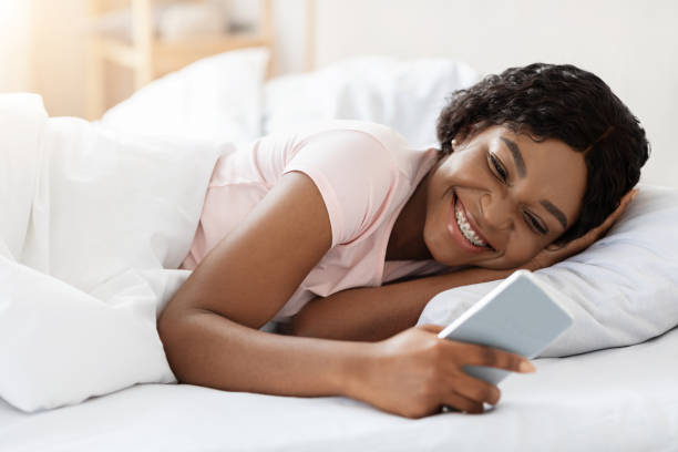 istock Happy young black woman using smartphone in bed 1295752837