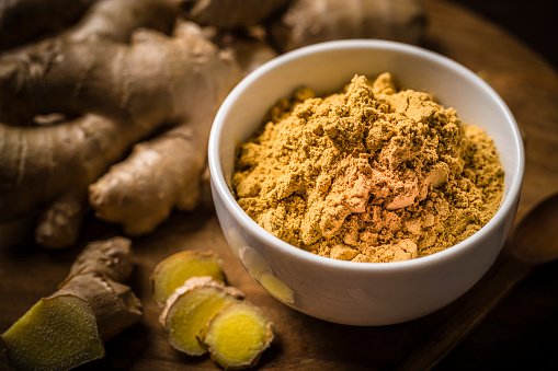Top view of a white bowl full of ground ginger spice surrounded by a fresh ginger root. Objects are on a rustic wooden table. predominant color is brown. Low key DSLR photo taken with Canon EOS 6D Mark II and Canon EF 100 mm f/ 2.8