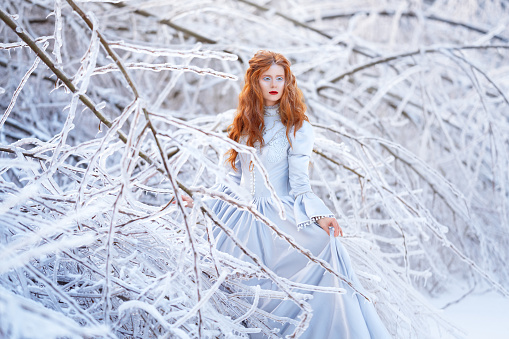 Young redhead woman, a princess, walks in a winter forest in a blue dress. Frost and snow on the trees.