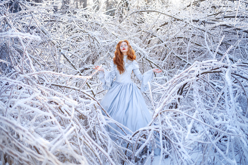 Young redhead woman, a princess, walks in a winter forest in a blue dress. Frost and snow on the trees.