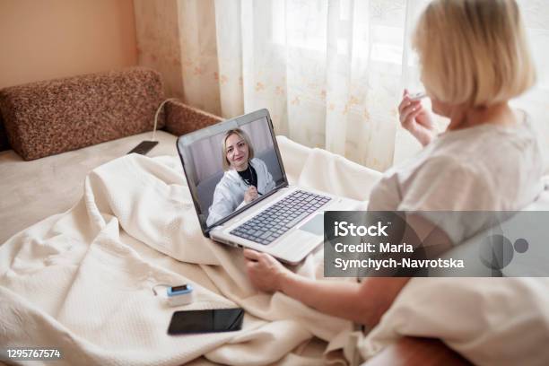 Old Woman In Bed Looking At Screen Of Laptop And Consulting With A Doctor Online At Home Telehealth Stock Photo - Download Image Now