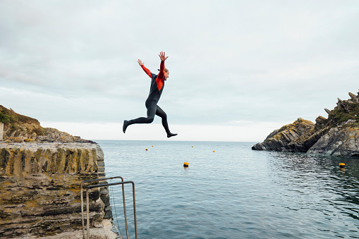 A side view of young man jumping into the harbor at Polperro, Cornwall. He is wearing a wetsuit.