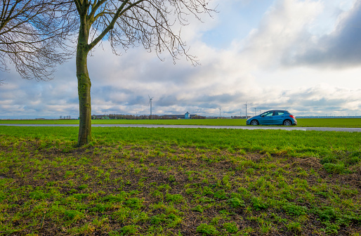 Car parked on a road along an agricultural field in the countryside under a blue cloudy sky in sunlight in winter, Almere, Flevoland, The Netherlands, January 8, 2021