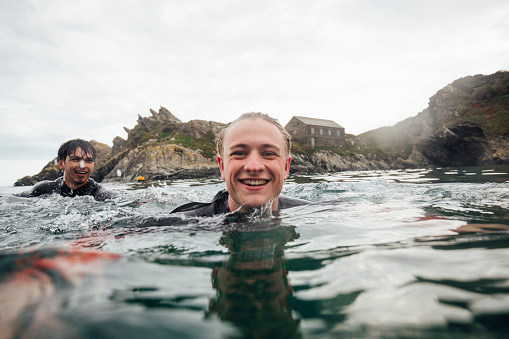A selfie shot of two young men swimming in the harbour water at Polperro, Cornwall. One is looking at the camera and smiling while the other one is swimming in the sea.