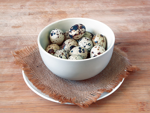 The quail eggs and a chicken egg in the ceramic bowl on the jute rug and ceramic plate on the wooden table. Closeup.