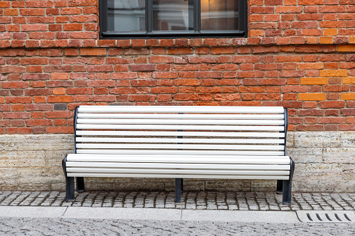 Lonely empty white wooden bench with old brown brick wall and black window on background.