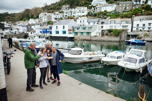 A group of male and female friends standing on a footpath next to the harbour in Polperro, Cornwall. They are sharing a tray of chips together.