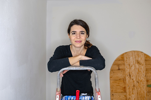 Smiling young woman on a ladder looking at the camera. House works, painting, renovation, handy woman concept.