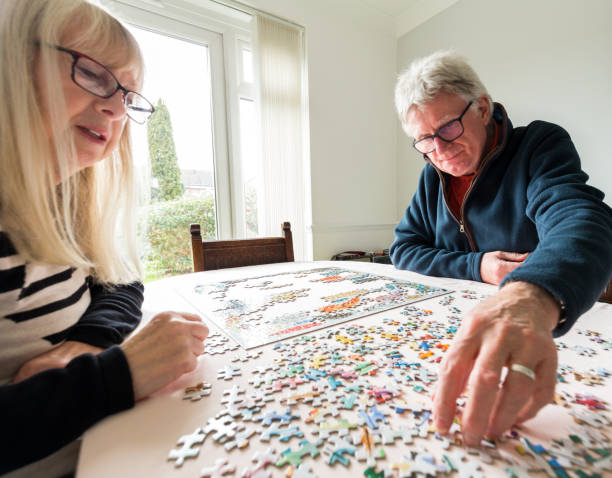 Color image depicting a senior caucasian couple, in their 60s and 70s, doing a jigsaw puzzle together at home during the coronavirus (Covid-19) pandemic. The couple are both wearing spectacles and are concentrating on completing the puzzle together. Room for copy space.