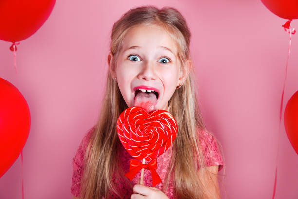 バレンタインデー。ピンクの背景にハート型のロリポップと赤いドレスを着た小さなかわいい女の子 - child valentines day candy eating ストックフォトと画像