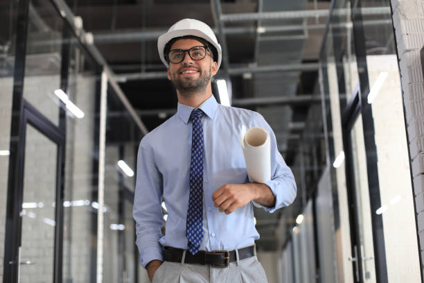 istock Confident young business man in hardhat holding blueprint and looking away while standing indoors. 1295790646