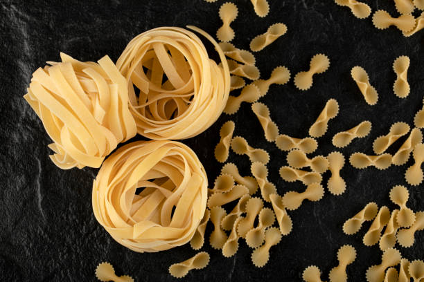 istock Raw tagliatelle nests and bow shaped pasta on black background 1295791468
