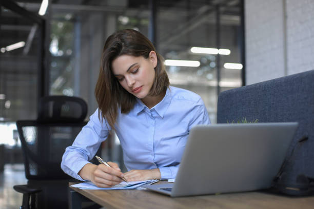 istock Smiling pretty woman sits at table. Happy entrepreneur writes notes to clients. 1295792289