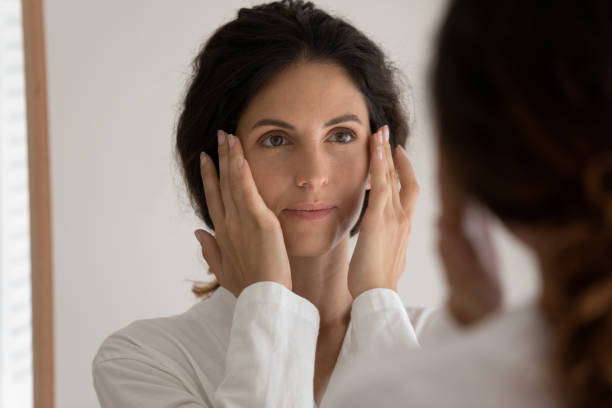 istock Young beautiful woman look in mirror massaging face 1295795447