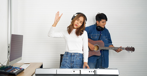 Young asian woman sings while playing an electric keyboard in front of white walls. Her boyfriend played the guitar together. Musicians producing music in professional recording studio.