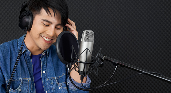 Musician producing music in professional recording studio. Young asian man with headphone singing in front of black soundproof wall.