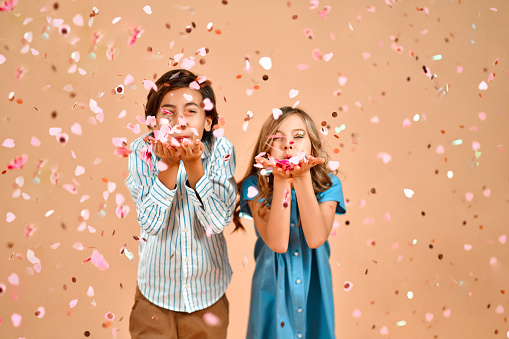 A cute boy in a striped shirt and a cute girl with curly hair in a blue dress are blowing confetti from their palms isolated on a gentle peach background.Valentine's Day.