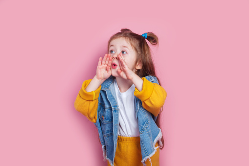 cute little girl with hands by mouth shouting on pink studio background