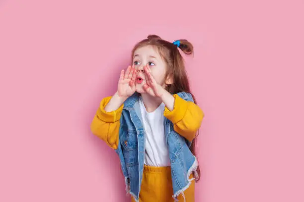 Photo of cute little girl with hands by mouth shouting on pink background