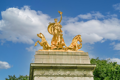 Close-up of the a sculpture group of gilded bronze figures of The USS Maine National Monument at the Merchant's Gate entrance to Central Park, Manhattan, New York, USA. The monument was cast on September 1, 1912 and dedicated on May 30, 1913 to the men killed aboard USS Maine (ACR-1) when the ship exploded in Havana harbor. Green trees and blue sky with clouds are in background.