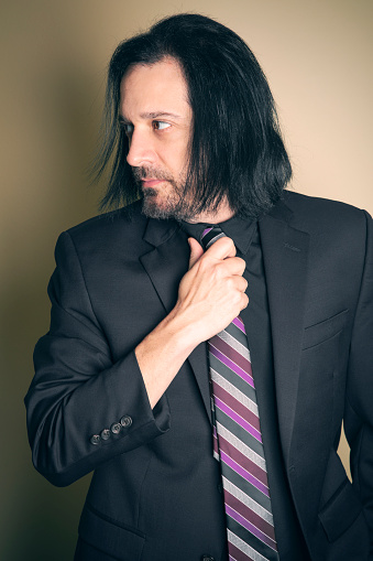 A handsome man with long hair in a suit looking to the side and adjusting his tie.