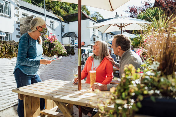 What Can I Get You? A couple sitting at a wooden table in a beer garden in Polperro, Cornwall, drinking alcoholic drinks. A waitress is taking their order at their table using a digital tablet, she is wearing a protective face mask to reduce the spread of Coronavirus during the COVID-19 pandemic. outdoor dining photos stock pictures, royalty-free photos & images