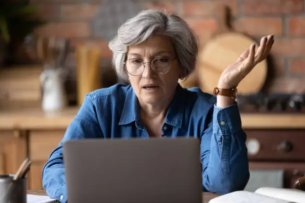 Confused middle aged 60s woman in glasses looking at computer screen, stressed of getting message with bad news. Unhappy mature lady feeling nervous about bad laptop work or poor internet connection.