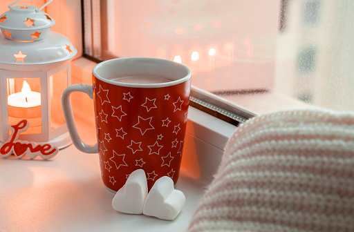 Cozy winter still life with red mug with hot drink, two white heart-shaped marshmallow, candle and scarf on the window. Valentine's, Christmas concept. Cute atmosphere image.
