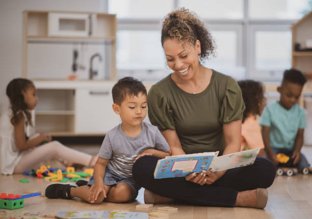 Reading with my teacher A preschool boy of Hispanic ethnicity sits next to his teacher as she is reading him a book on the classroom floor. montessori education photos stock pictures, royalty-free photos & images